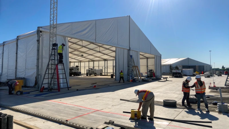 Workers assembling a large warehouse tent at a construction site under a blue sky.